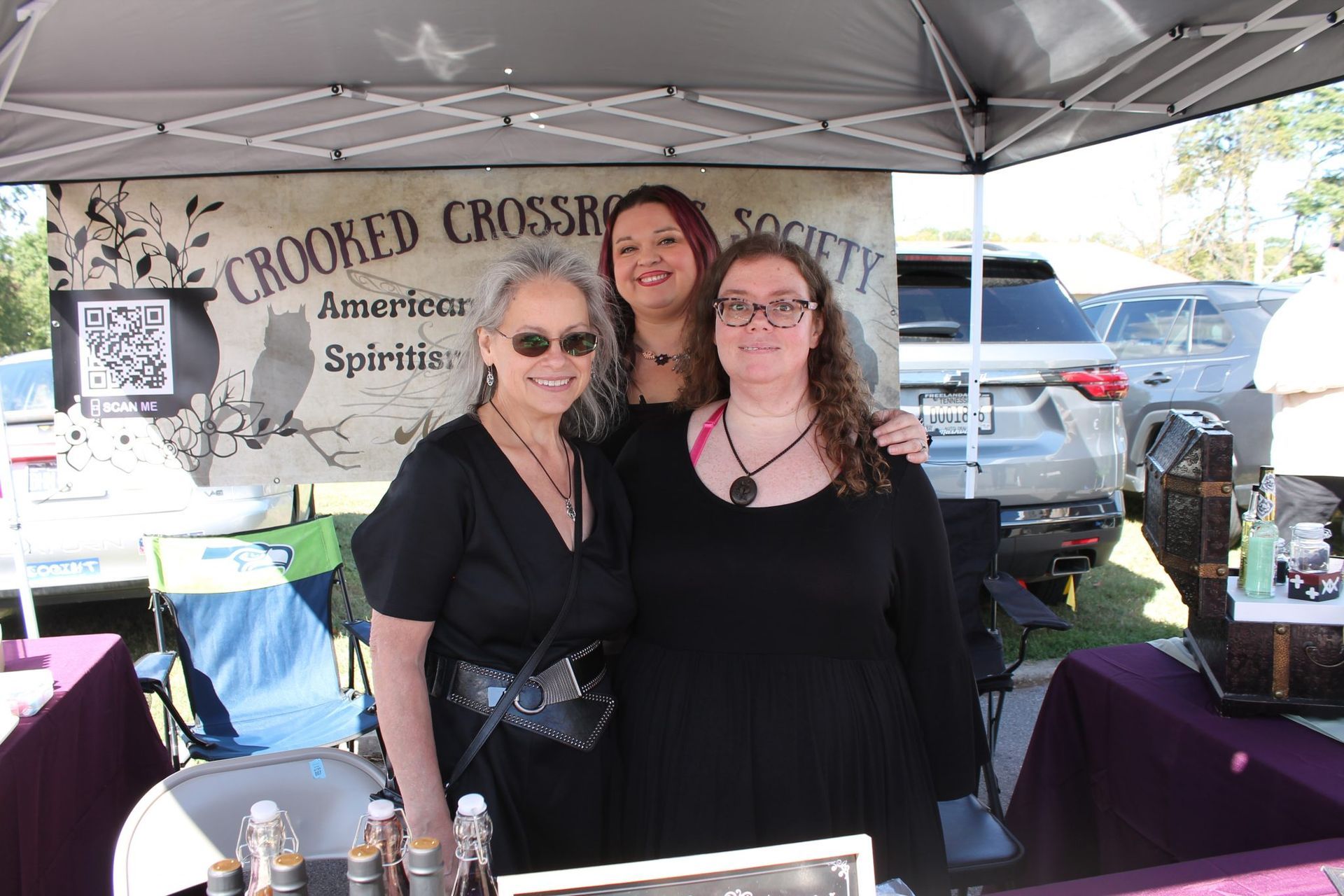 Three women posing under a canopy at a Crooked Cross Brewing Society booth with bottles on display