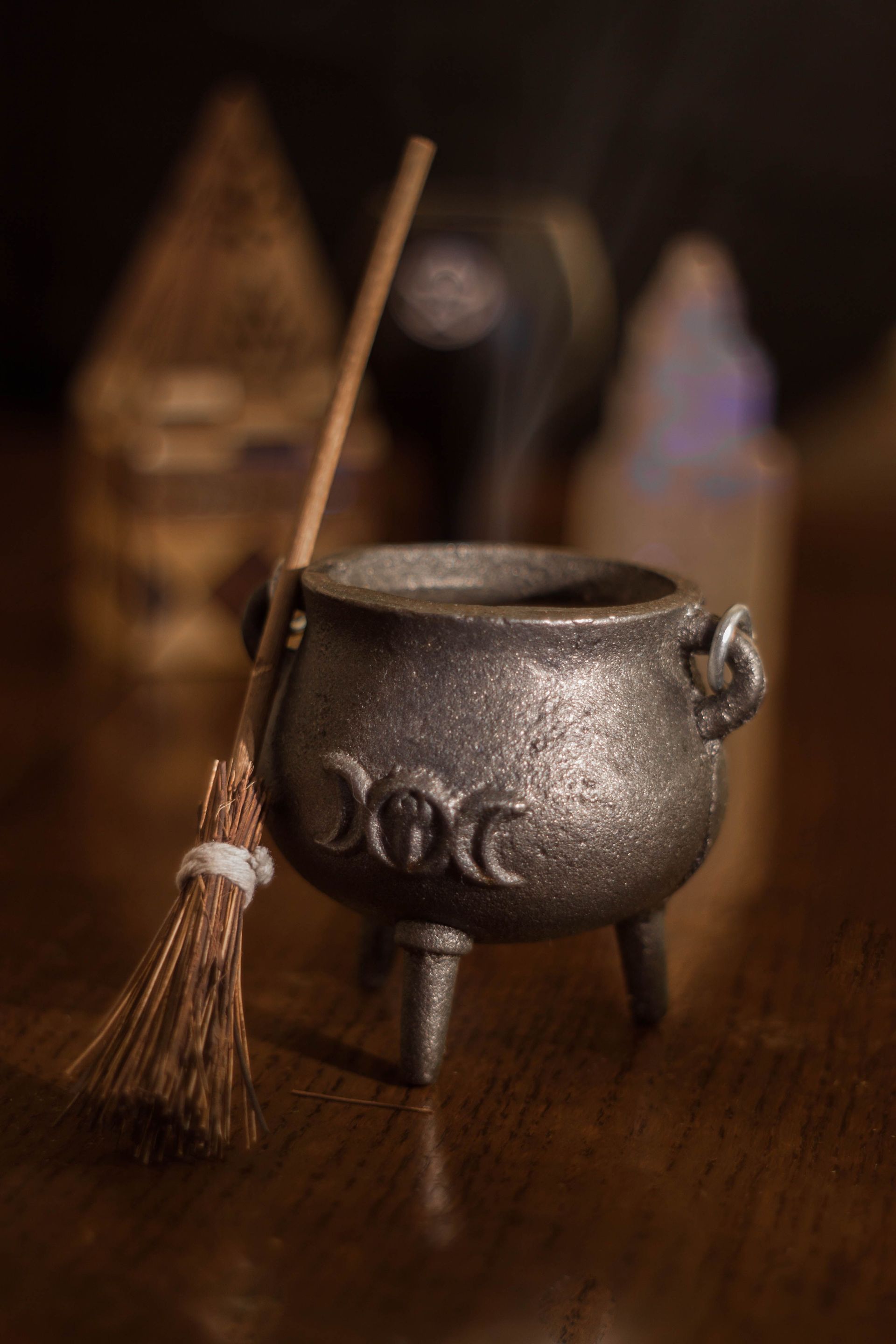 Small silver cauldron with a spoon and broom on a wooden table, with blurred spooky decor in back