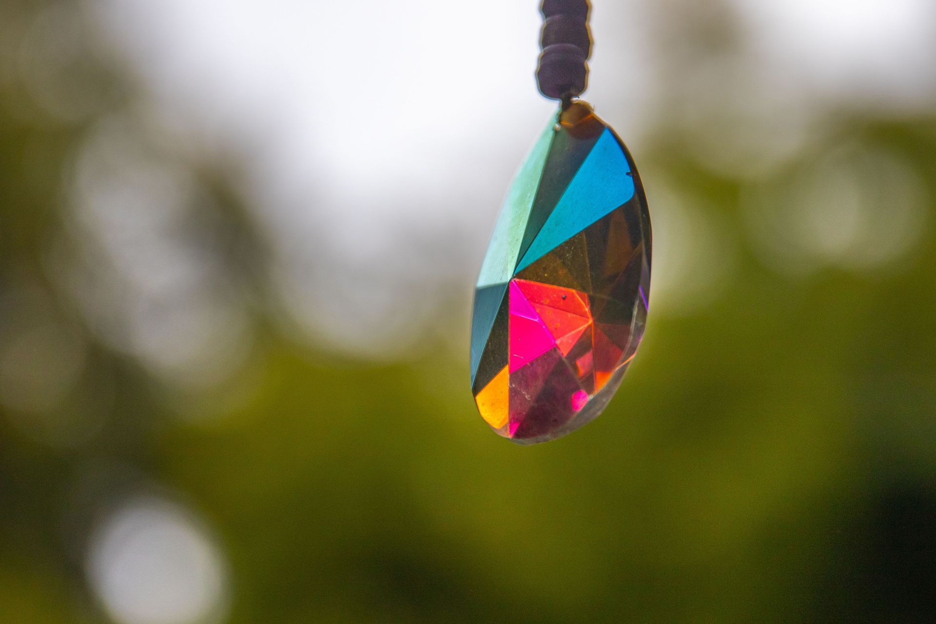 Faceted teardrop crystal pendant hanging outdoors, reflecting blue, pink, and gold light