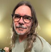 Portrait of a person with long gray hair, glasses, and a mustache, against a softly lit background