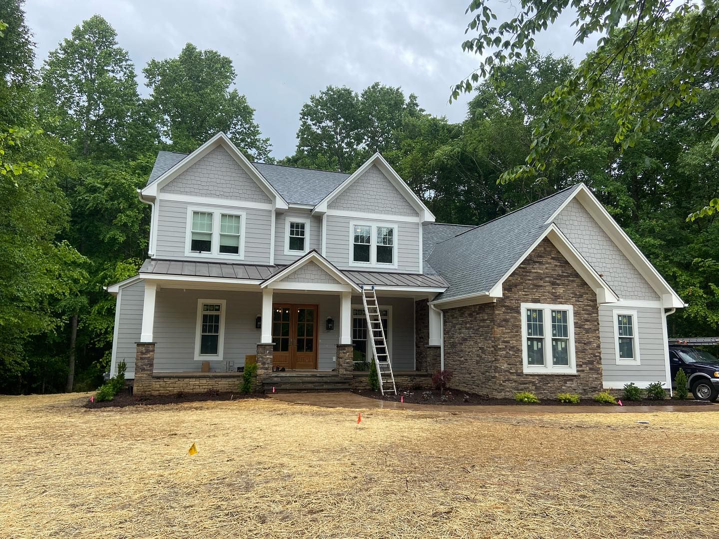 Two-story house with gray siding, stone accents, and a ladder in front, set against a backdrop of trees.