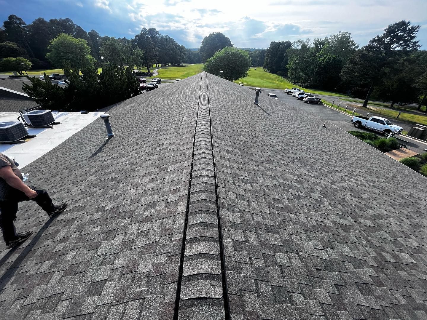 Man on a gray shingle roof with a central ridge, trees and sky in the background.
