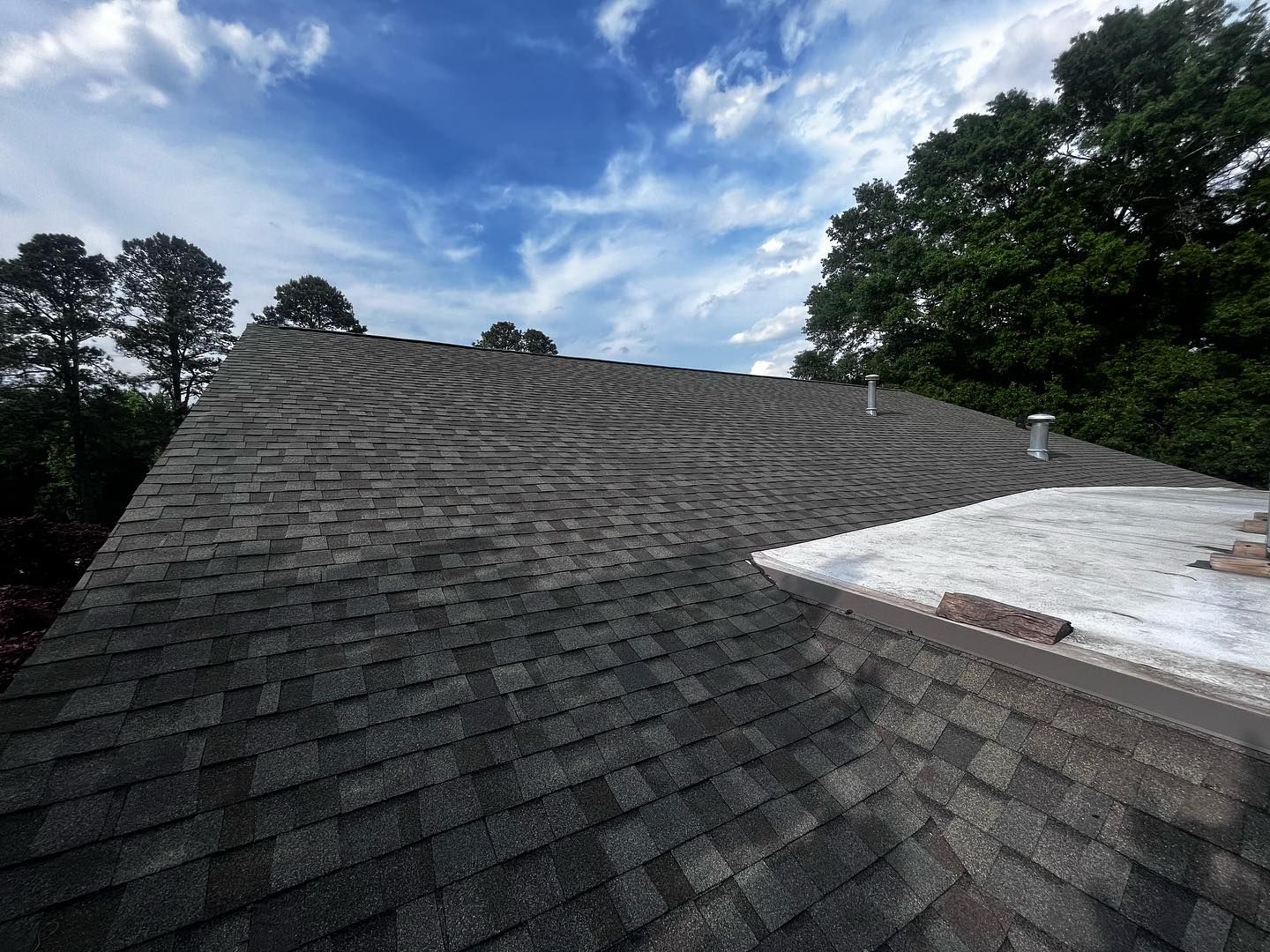 commercial roofers mechanicsville va Roof covered in dark shingles under a cloudy blue sky with trees in the background.