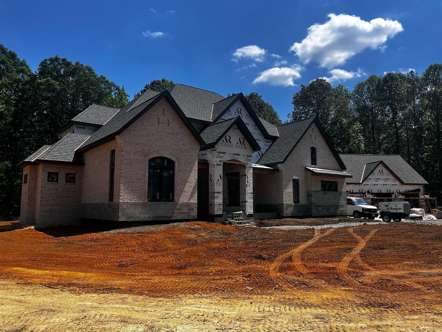 New house under construction with brick exterior and dark roof, on a dirt lot with trees in background.