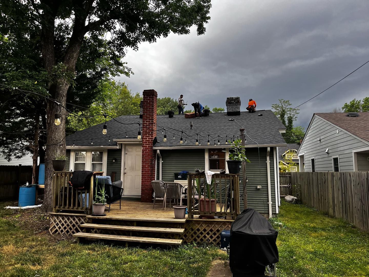 roof repairs near me Workers on a roof, replacing shingles. Green house, brick chimney, backyard setting, overcast sky.