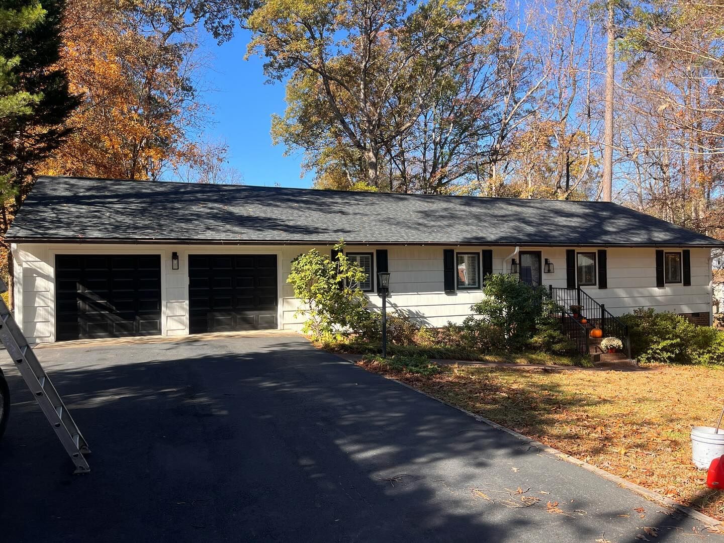 A one-story white house with a black roof and garage doors, set against autumn trees and a clear blue sky.