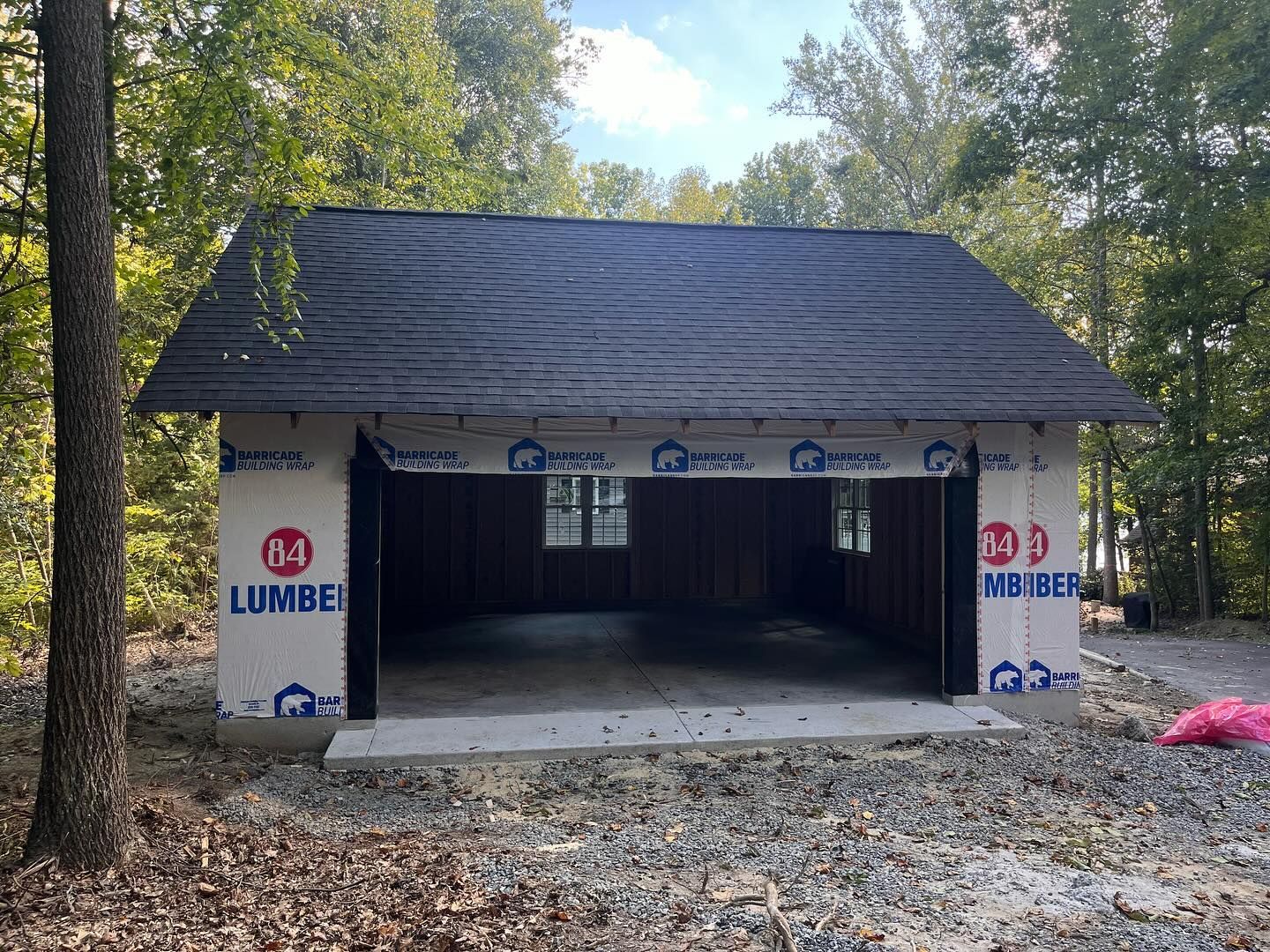 Unfinished garage with dark roof, open front, and paper wrap on the walls, set in a wooded area.