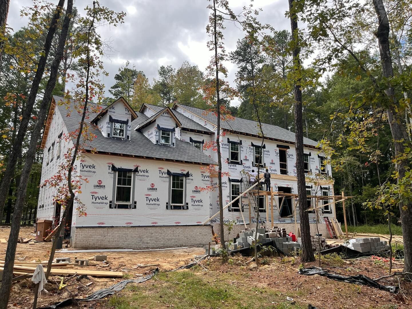House under construction in a wooded area, wrapped in white paper, with visible windows and scaffolding.