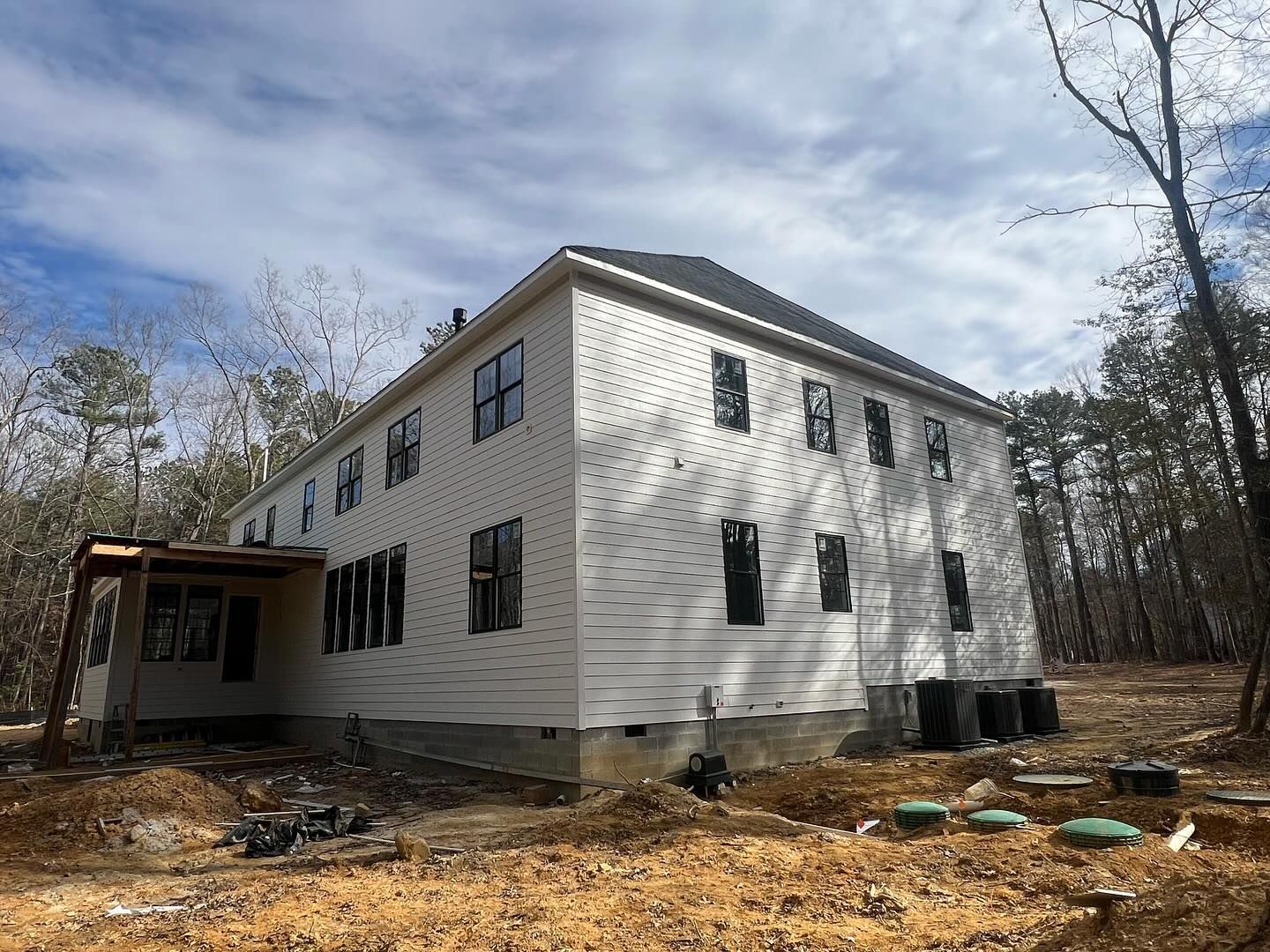 Two-story house under construction, white siding, black window frames, brown wooden porch, set in a wooded area.
