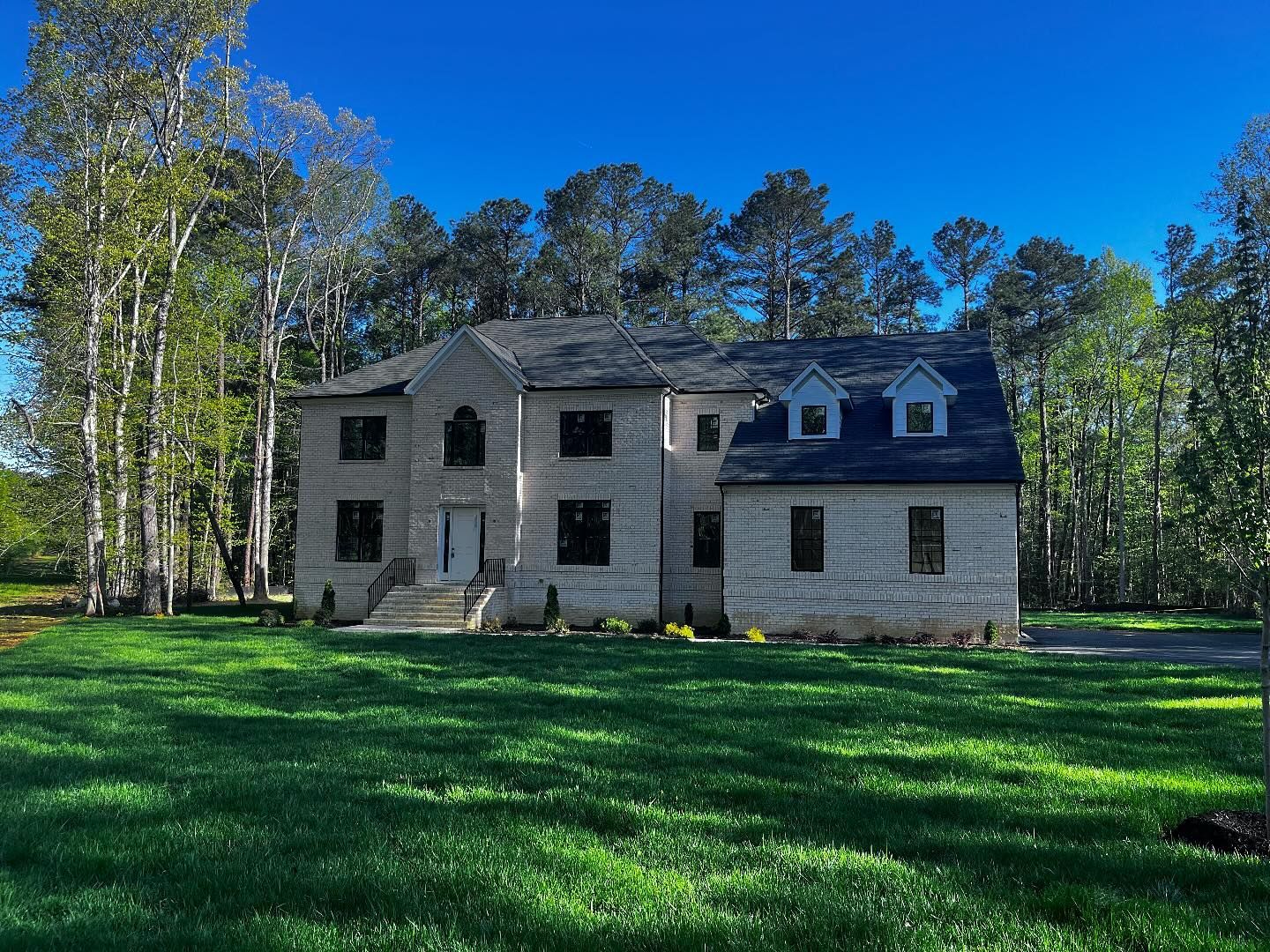 Two-story beige brick house with black windows and blue roof, surrounded by green grass and trees on a sunny day.