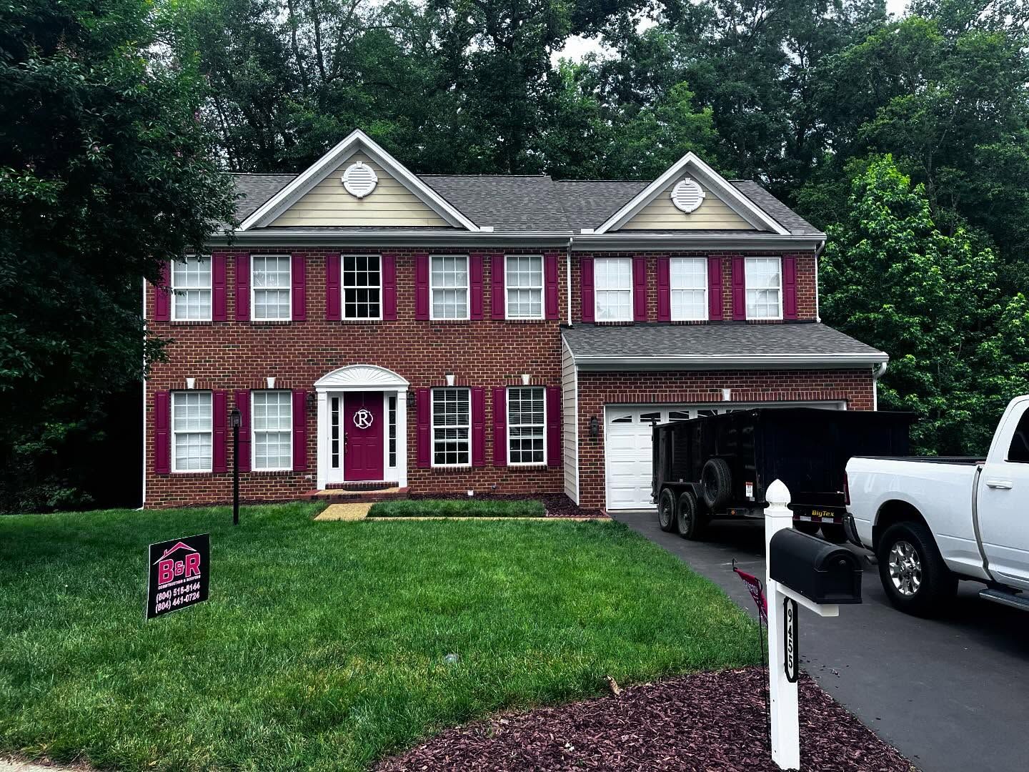 Brick house with red shutters, white trim, and a dark-colored trailer in the driveway.