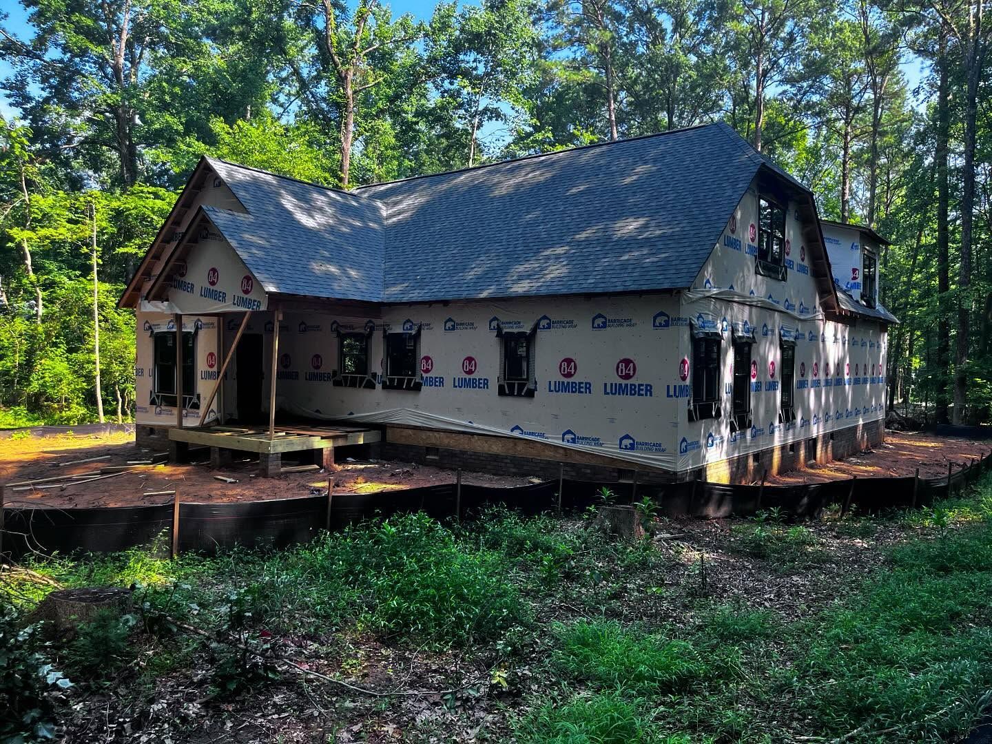 House under construction surrounded by trees and green grass. Gray roof, blue tarp, windows visible.