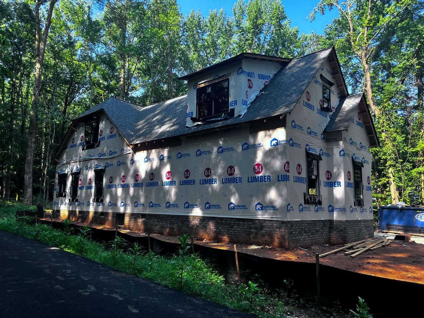 New house under construction, exterior view. Stone foundation, blue wrap, gray roof, surrounded by trees.