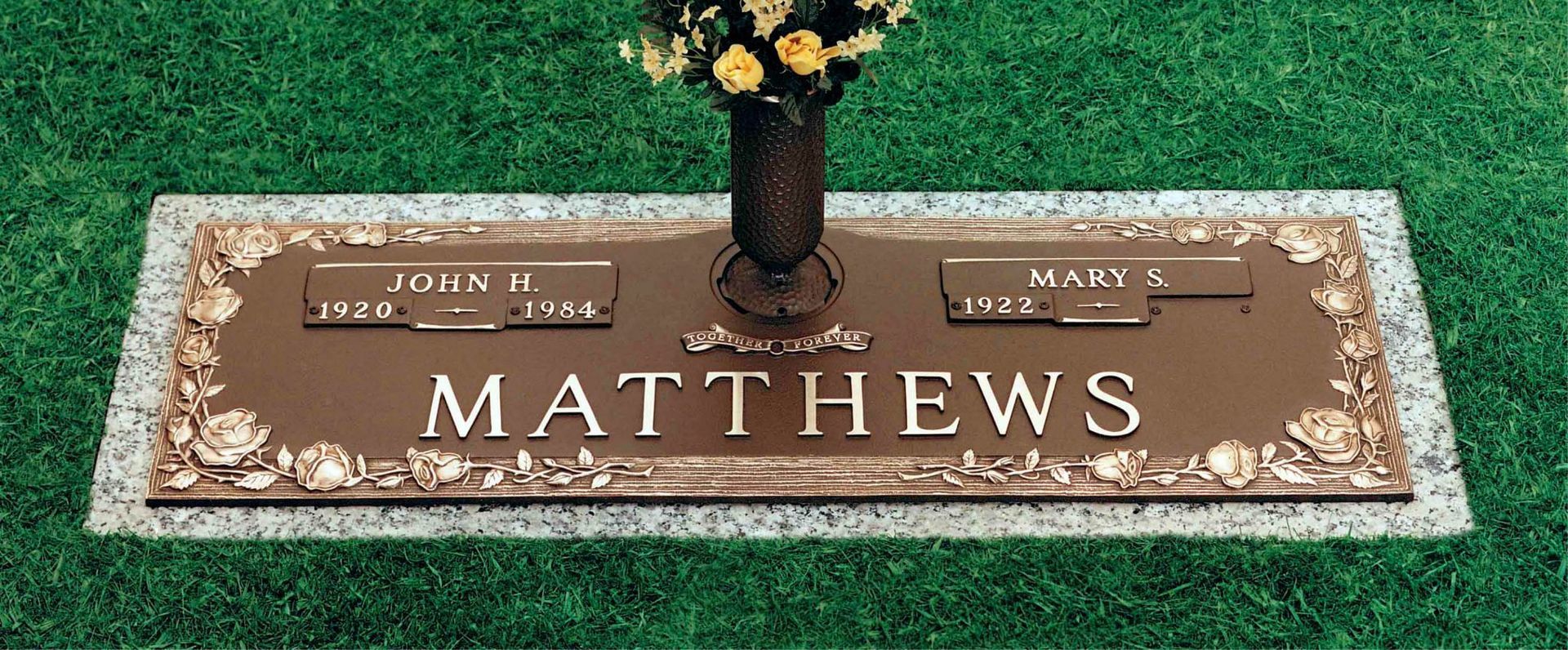 Bronze memorial grave marker with the name “Mathews” on green grass, topped with a vase of flowers