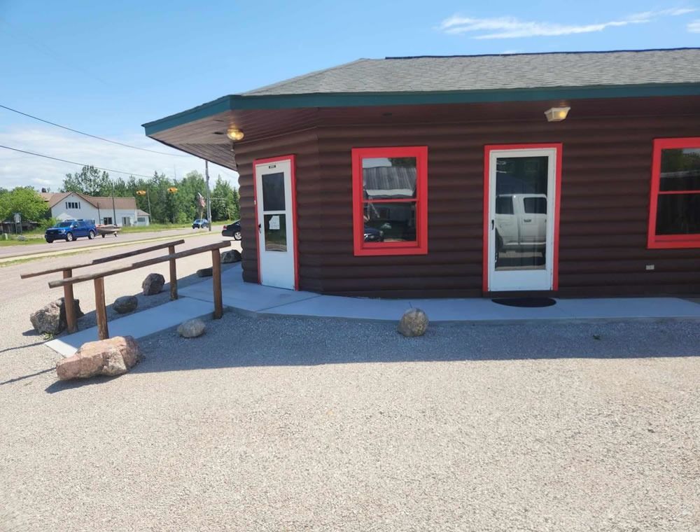 Small roadside building with red-trimmed windows, white door, and a wooden ramp on a gravel lot.