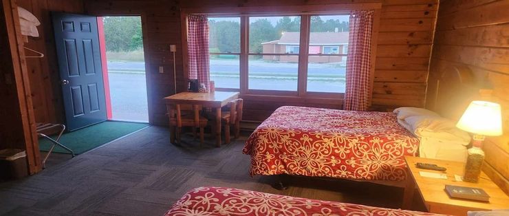 Rustic cabin bedroom with two beds, wooden walls, a table by windows, and an open door to outside.
