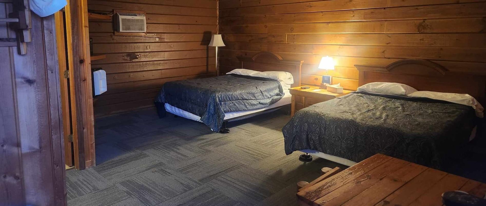Cozy wood-paneled cabin bedroom with two beds, a warm lamp, and a small table on a carpeted floor