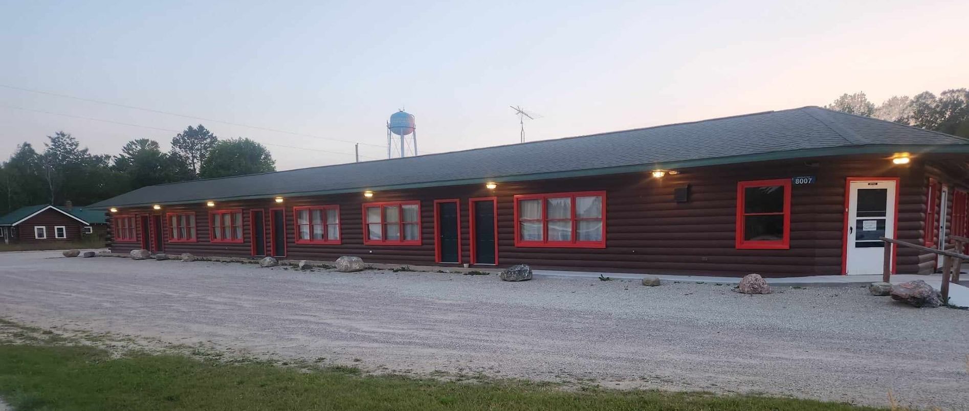 Long red motel-style building with lit windows along a gravel lot at dusk.