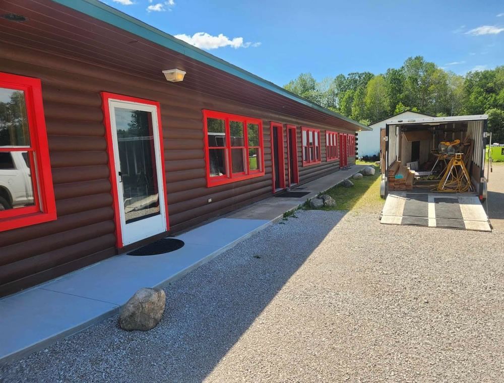 Red building with bright red windows and door beside a gravel driveway and open trailer in sunny daylight