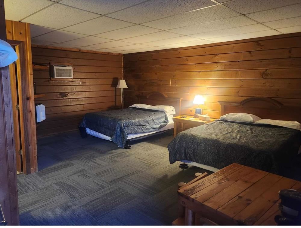 Rustic wood-paneled bedroom with two beds, bedside lamp, and a wooden table.