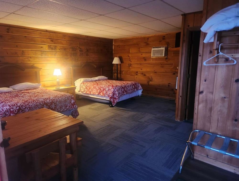 Wood-paneled motel room with two beds, warm lamp light, desk, and open closet wardrobe