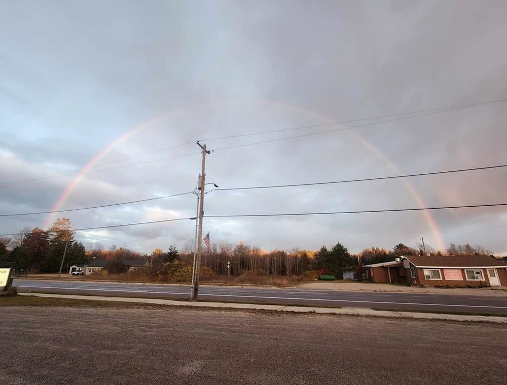 Rainbow over a rural roadside with power lines, trees, and a small building under cloudy sky