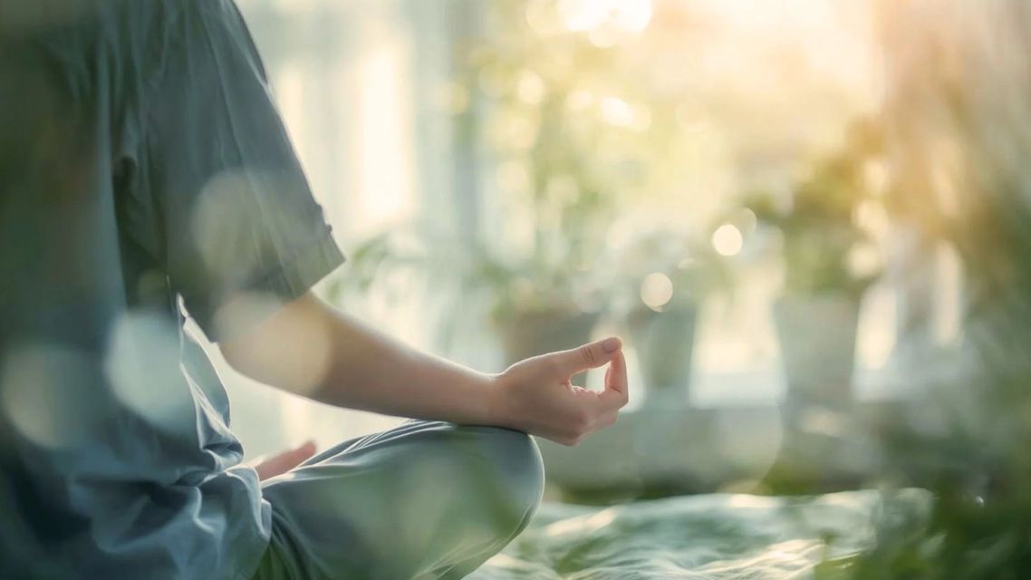 Person sitting cross-legged in a sunlit room, meditating with one hand resting on the knee.