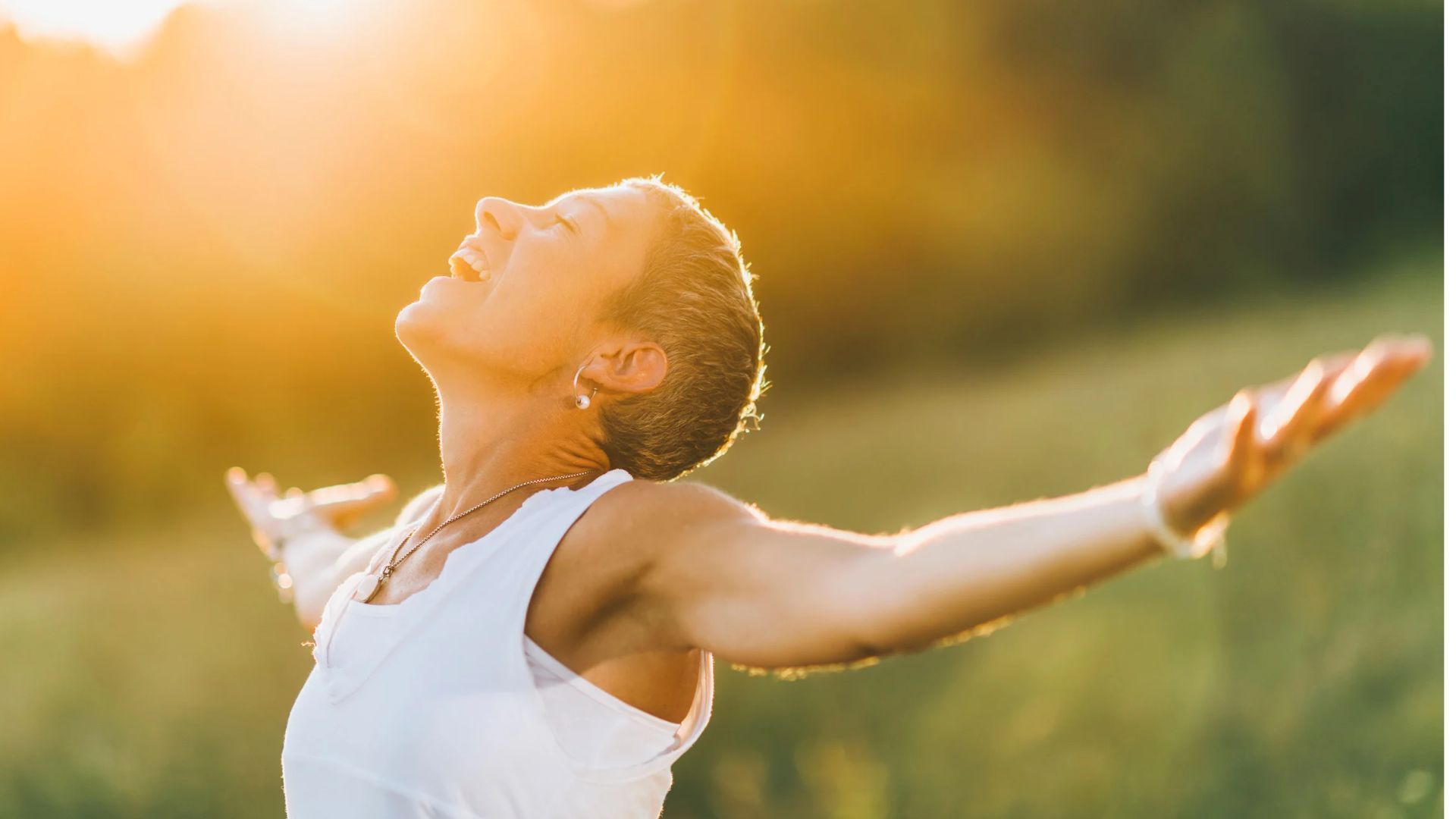 Person in a white top stands outdoors with arms outstretched, face lifted into warm sunlight.