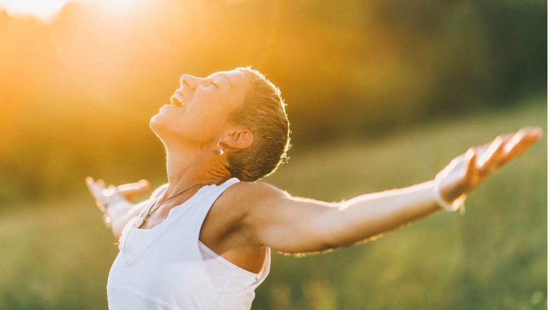 Person in a white top stands outdoors with arms outstretched, face lifted into warm sunlight.