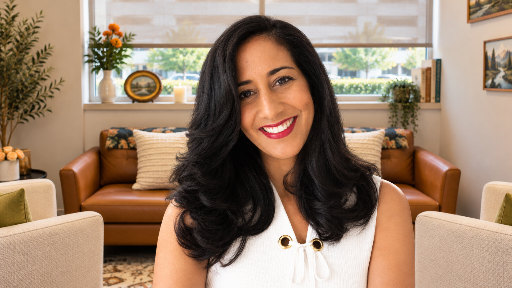 Smiling woman with long dark hair in a bright living room with tan chairs and potted plants