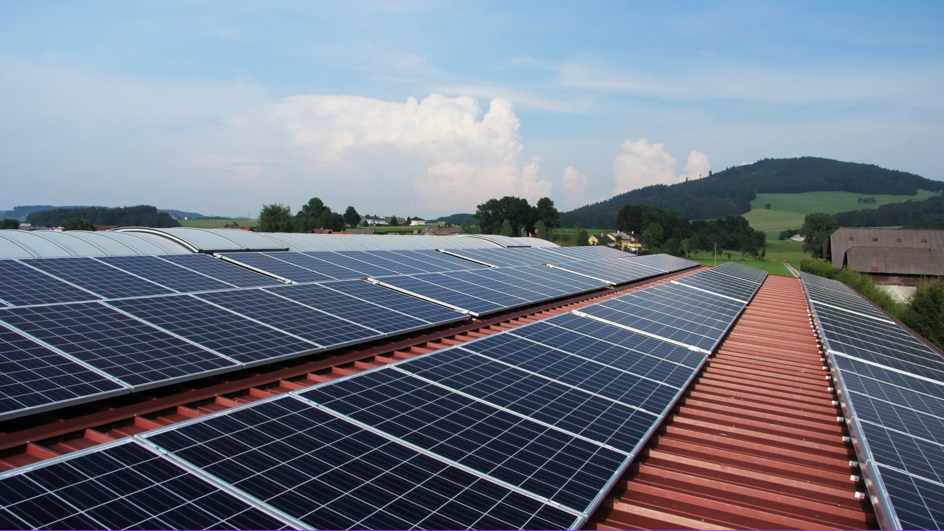 Panneaux solaires sur un toit en tuiles rouges, avec un paysage verdoyant et un ciel bleu en arrière-plan.