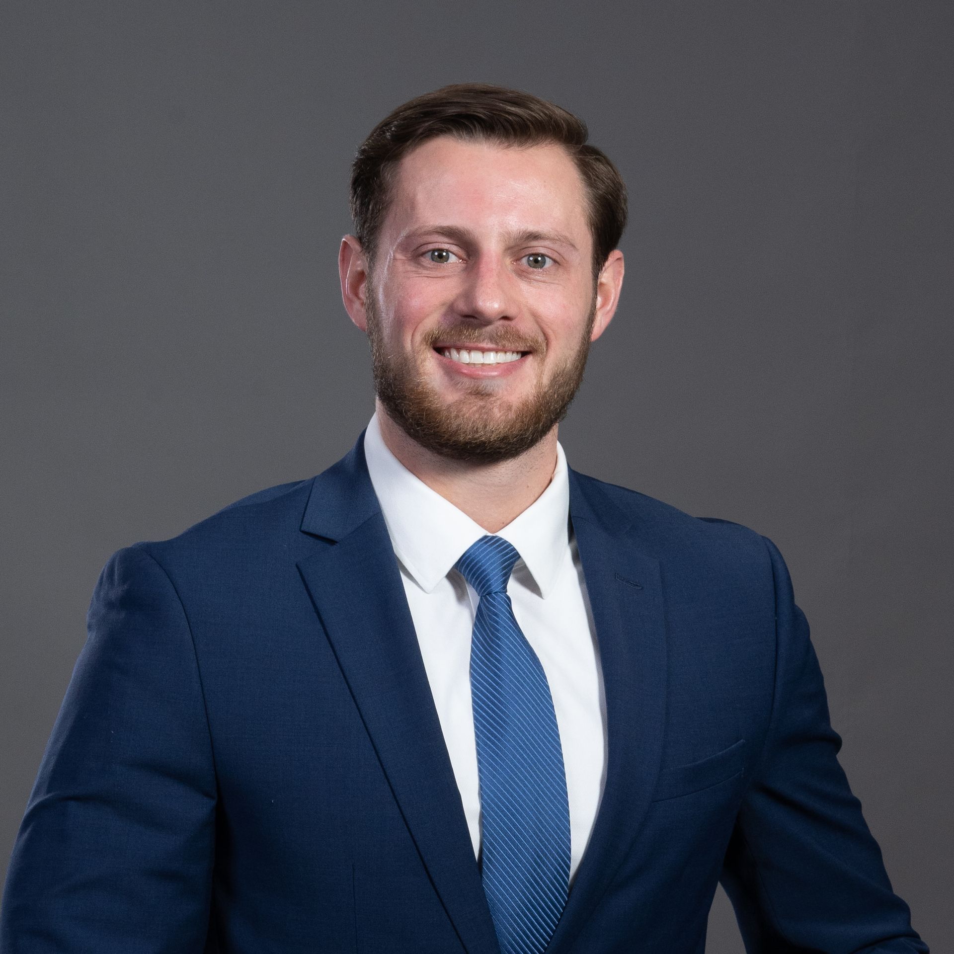 Portrait of a smiling man in a navy suit and blue tie against a gray background