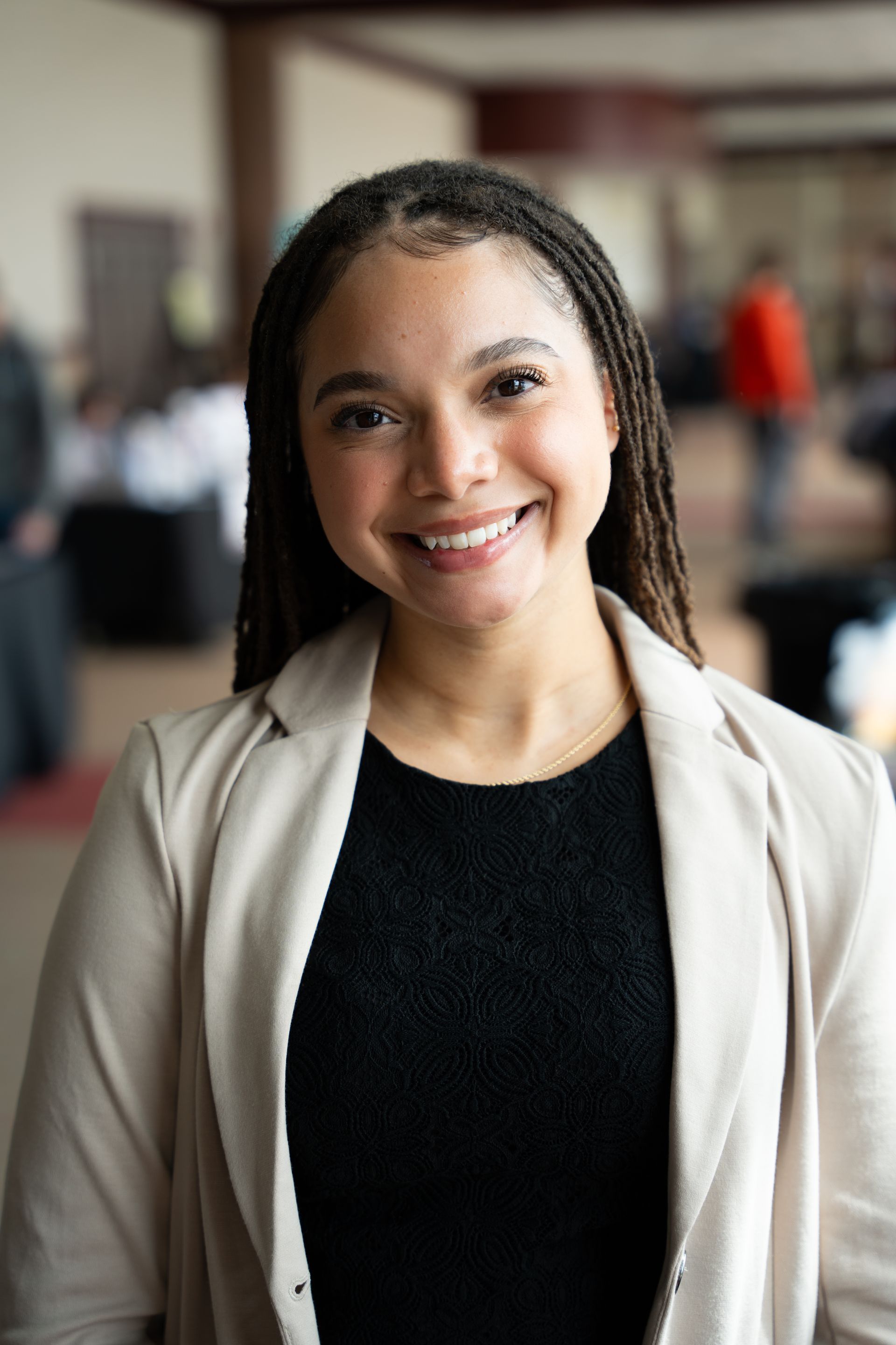 Smiling person in a beige blazer and black top standing indoors with a blurred office background
