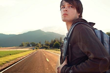 A young man with a backpack is standing on the side of a road.