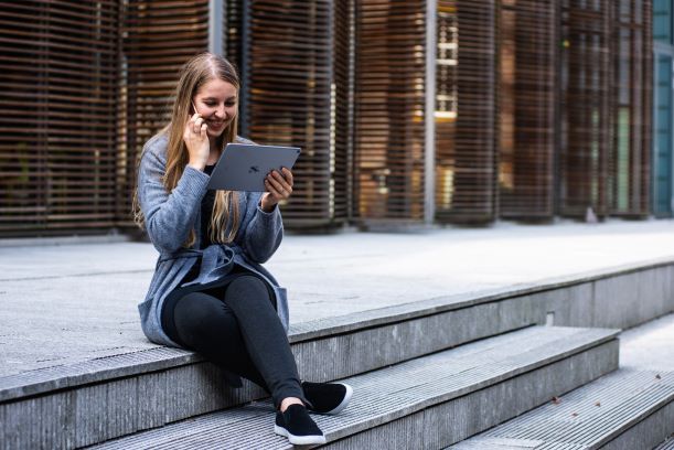 A woman is sitting on a set of stairs using a tablet and talking on a cell phone.