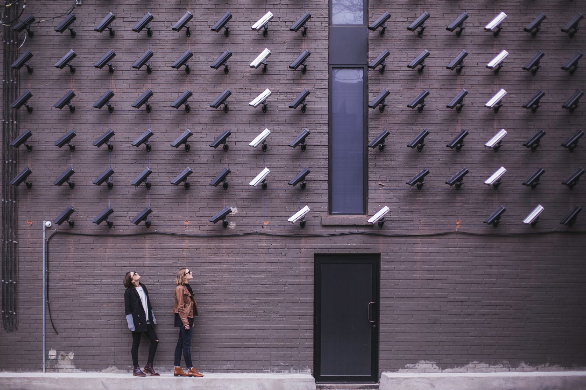 Two women are standing in front of a wall with a lot of cameras on it.