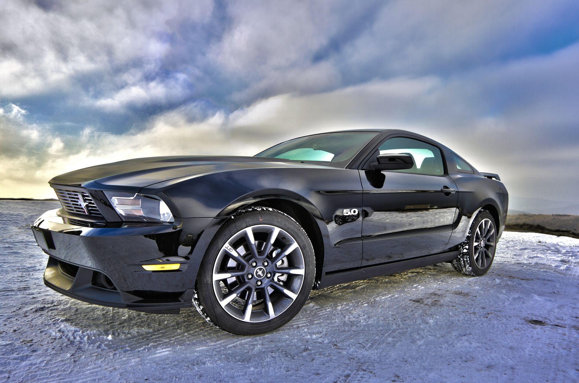 A black mustang is parked in the snow on a cloudy day.