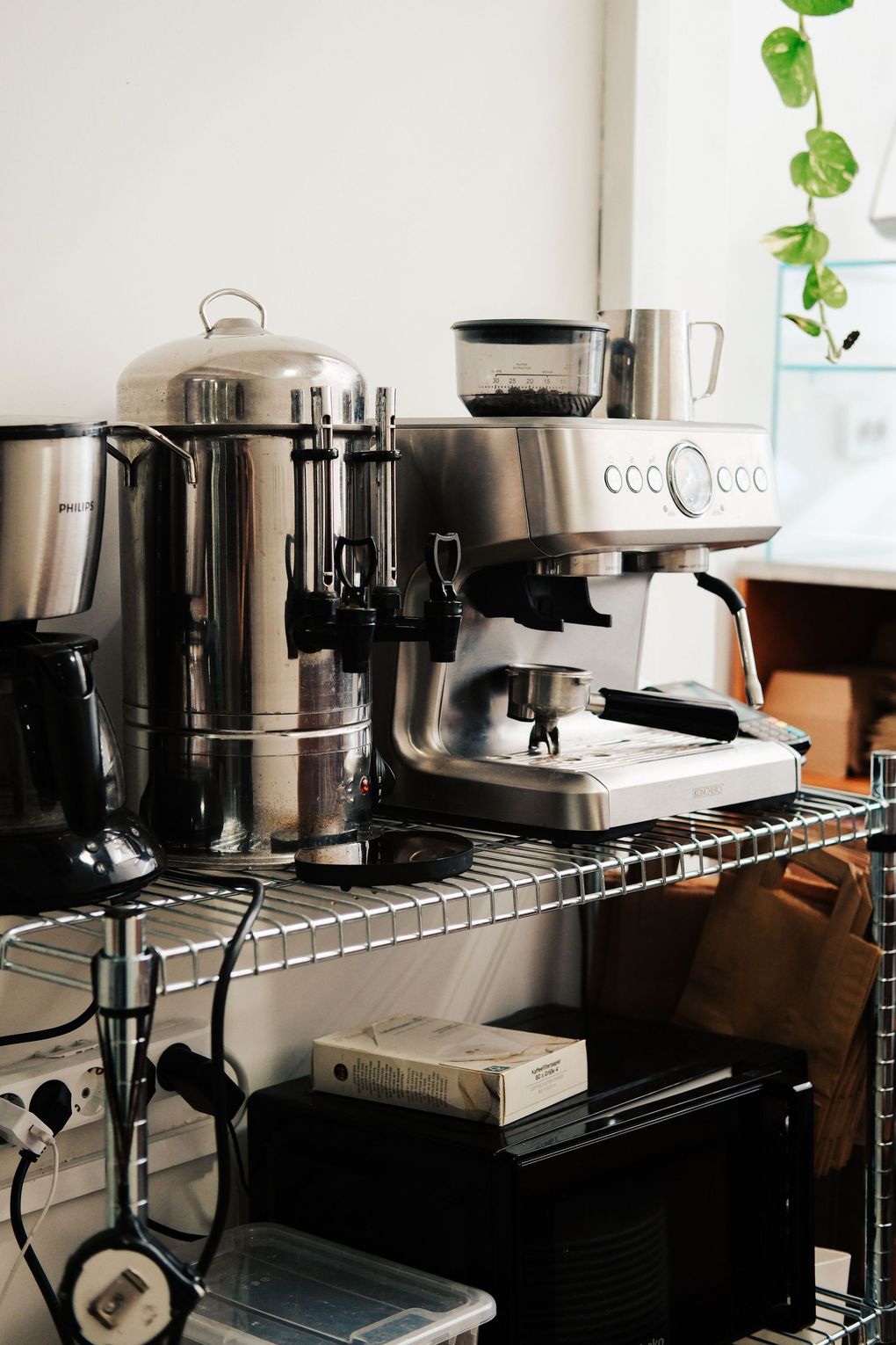 Coffee station with espresso machine, grinder, and cups on a metal shelf by a window with hanging plants