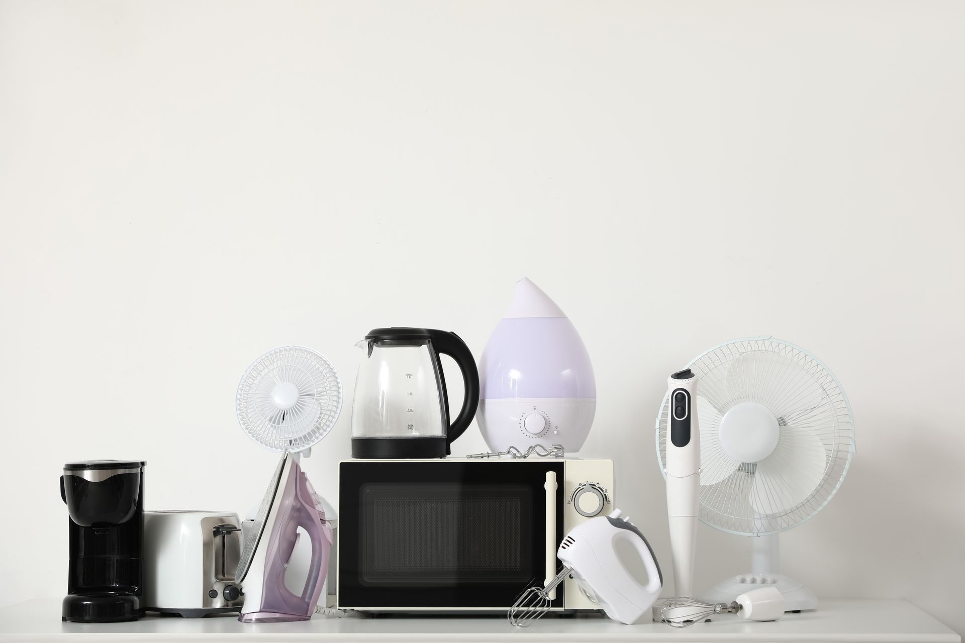 Assorted kitchen appliances lined up on a white shelf against a white wall.