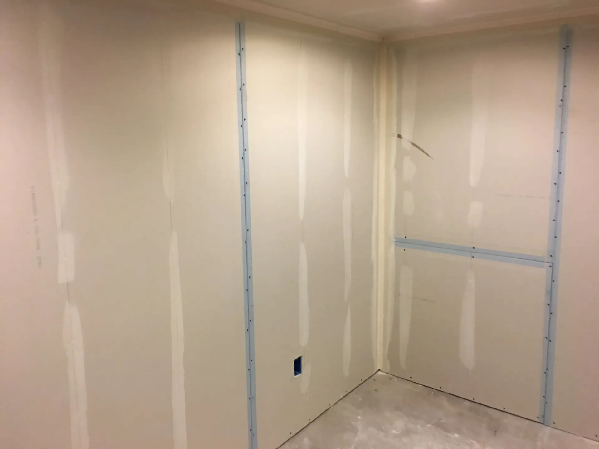 Empty beige room with unfinished drywall, metal studs, and exposed electrical outlet socket