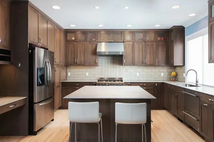 Modern kitchen with dark wood cabinets, stainless appliances, island, and two stools under bright recessed lights