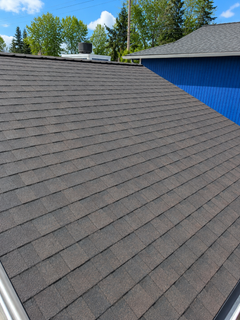 Gray shingled roof beside a blue house under a clear sky with trees in the background