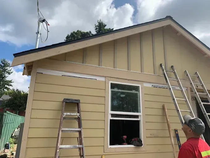 Two workers on ladders painting a beige house exterior under a cloudy sky.