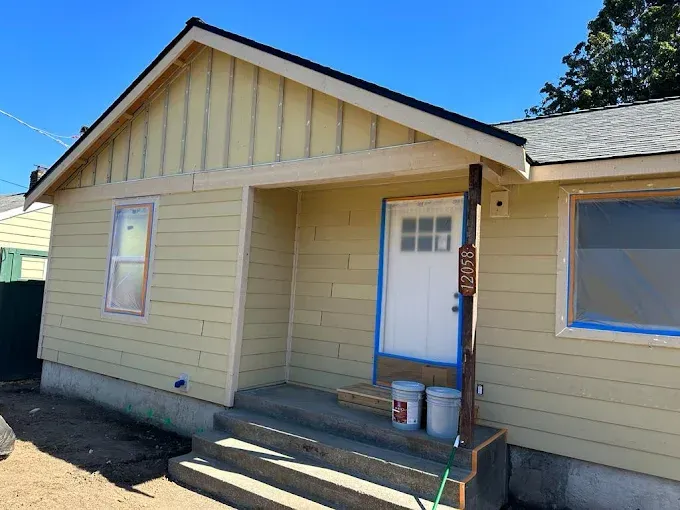 Front view of a small beige house with blue trim, a covered porch, and steps under a clear sky