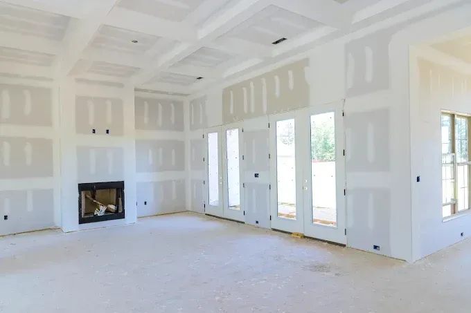 Bright unfinished living room with white drywall, fireplace, and large windows overlooking greenery