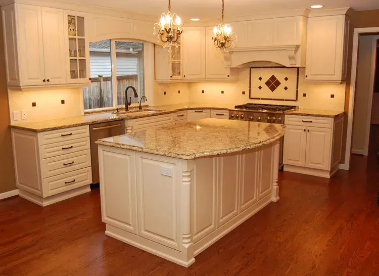 Bright white kitchen with granite island, wood floors, chandeliers, and a window over the sink.