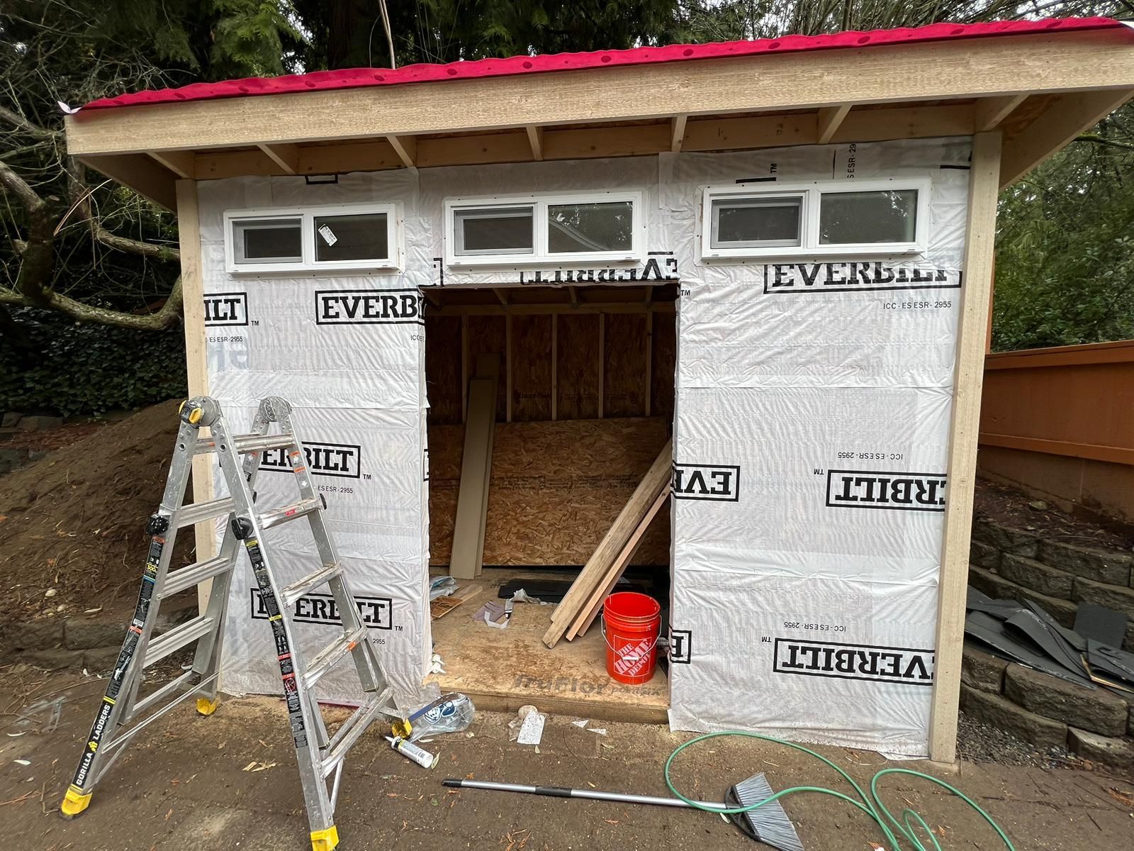 Small shed under construction with insulation, ladder, open doorway, and red bucket outside.