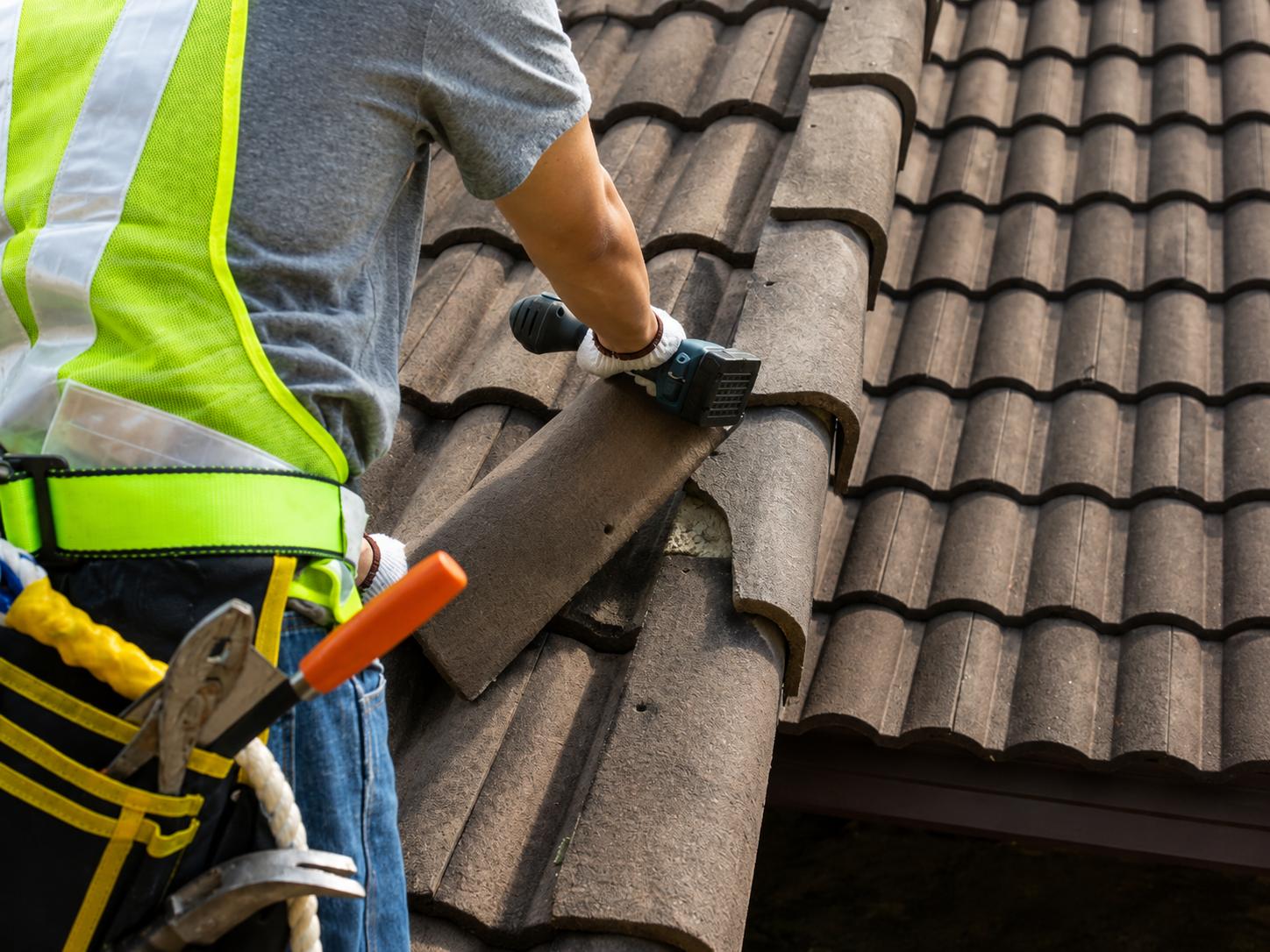Roofing worker lifting a brown shingle on a tiled roof, wearing a safety harness and gloves.