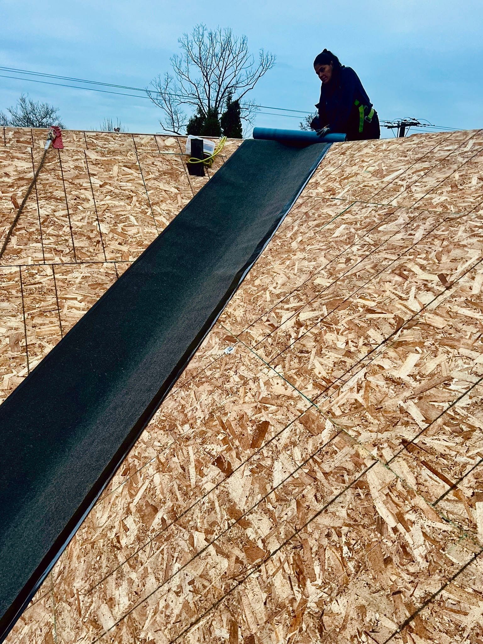 Worker on a roof applying underlayment along a ridge under a blue sky.