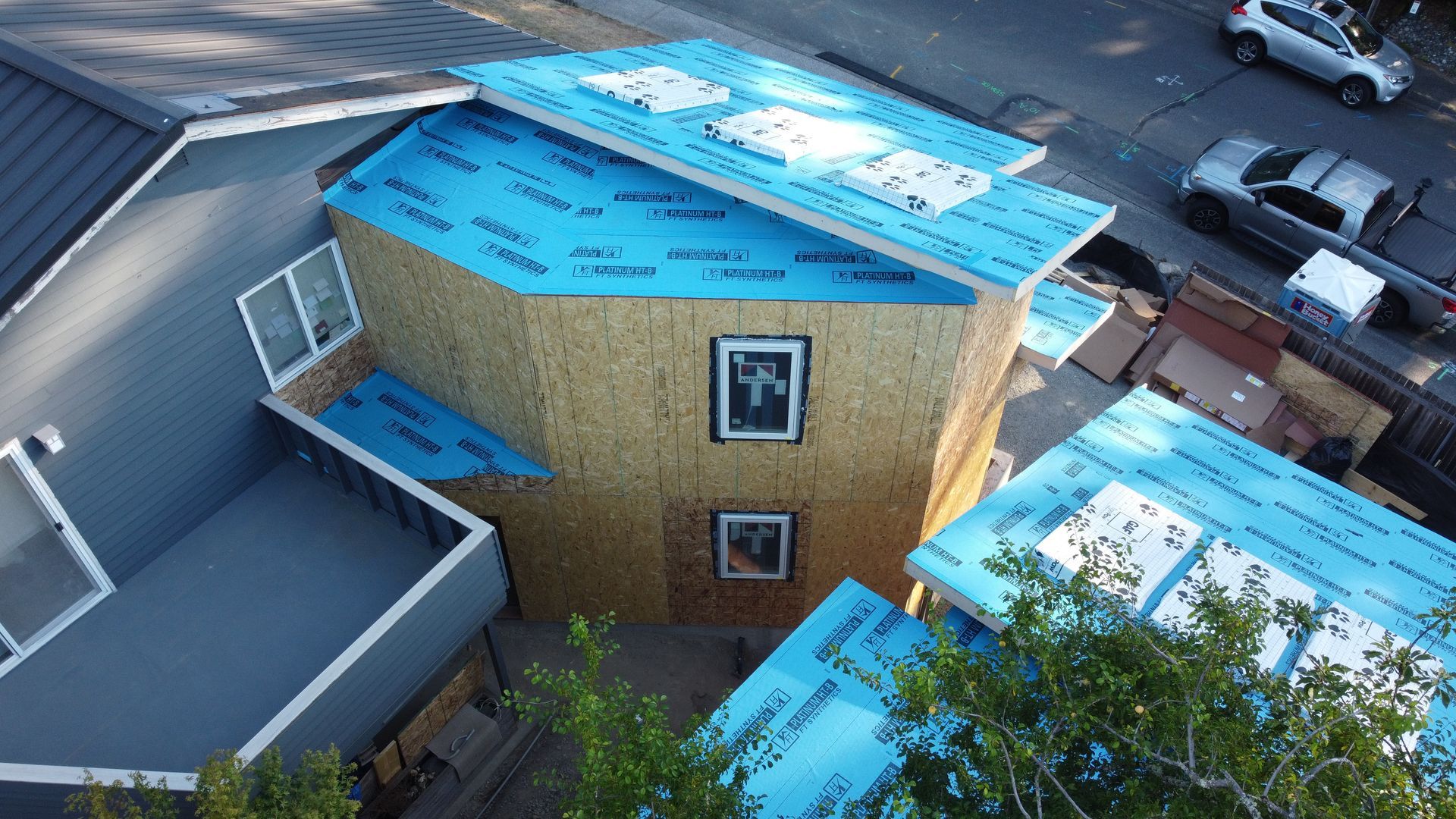 Aerial view of a house roof under construction with blue underlayment and a nearby street and parked cars